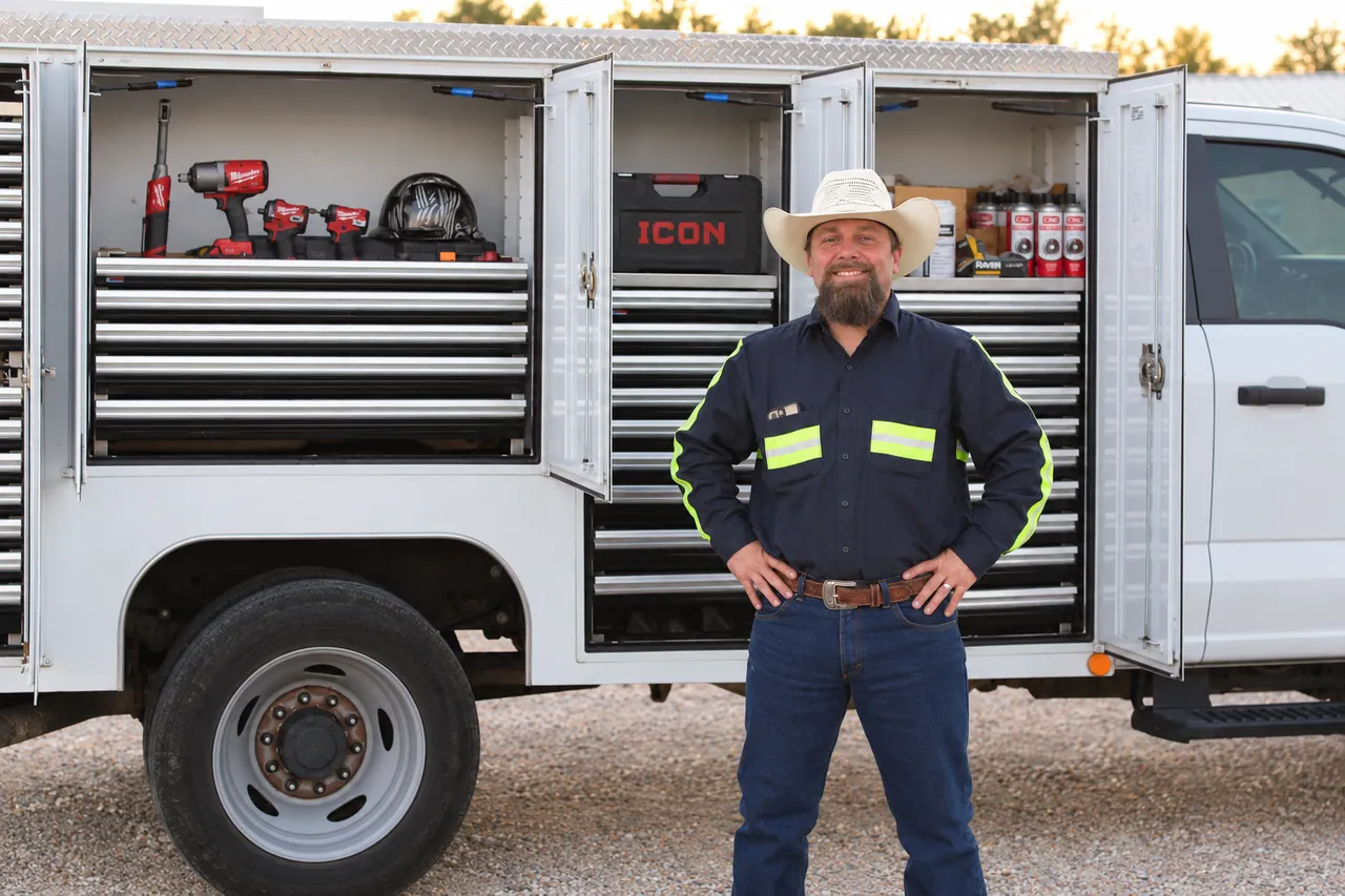 Kenneth Krickbaum in a cowboy hat and hi-vis work shirt, smiling with hands on hips in front of his service truck, with impact tools and a hard hat in the open compartment behind him.